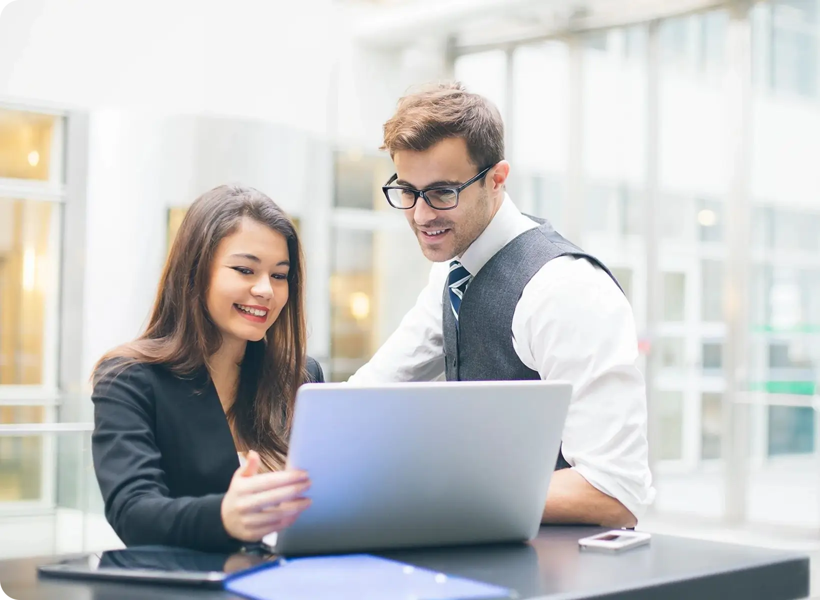 Two professionals are smiling and working together on a laptop in a modern office setting. One is seated, pointing at the screen, while the other stands beside, looking at the screen.