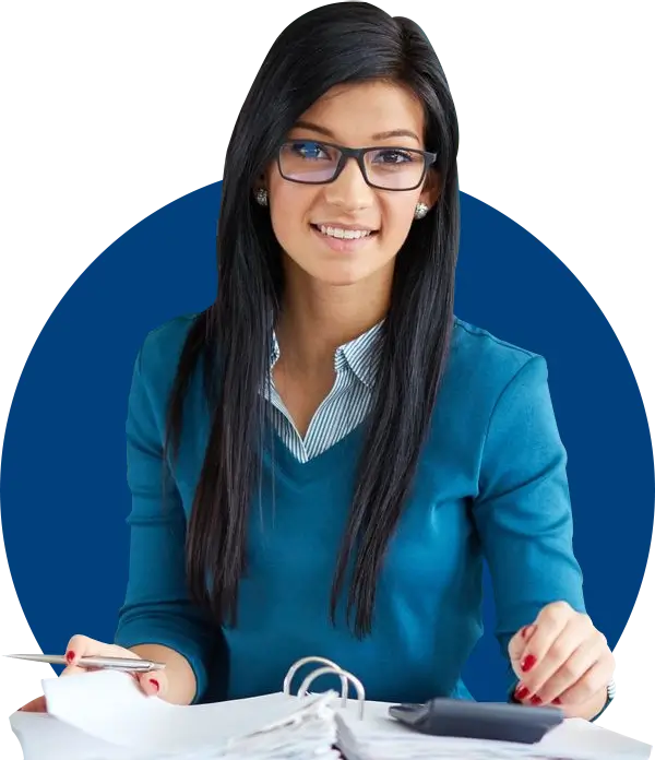 A woman with long dark hair and glasses sits at a desk, smiling, with open documents and a pen in hand. She is wearing a blue sweater.