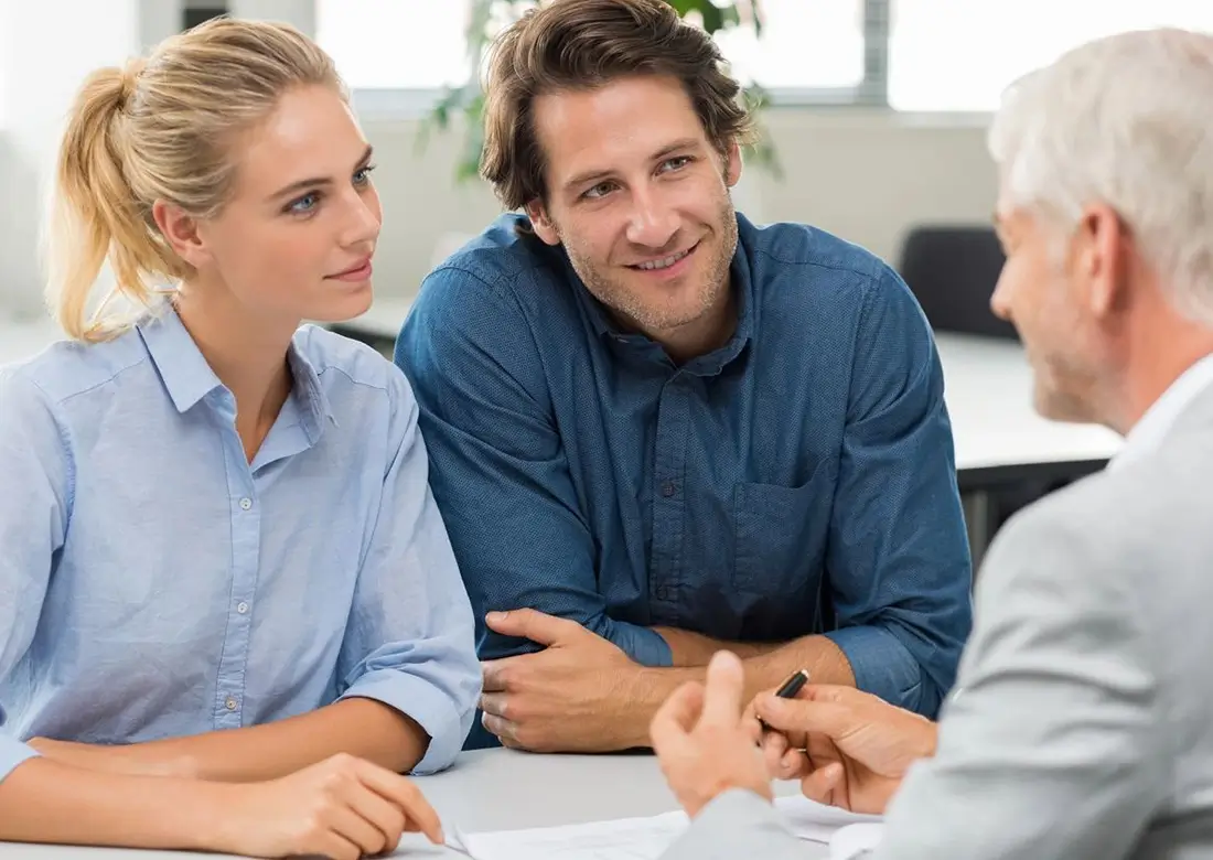 A young woman and man sit across a table from an older man in a suit. All three are engaged in conversation, with paperwork visible on the table.
