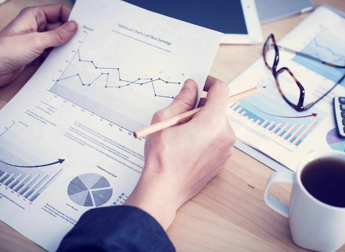 Person analyzing financial graphs and charts on paper while holding a pencil, with a tablet, glasses, calculator, and coffee cup on the desk.