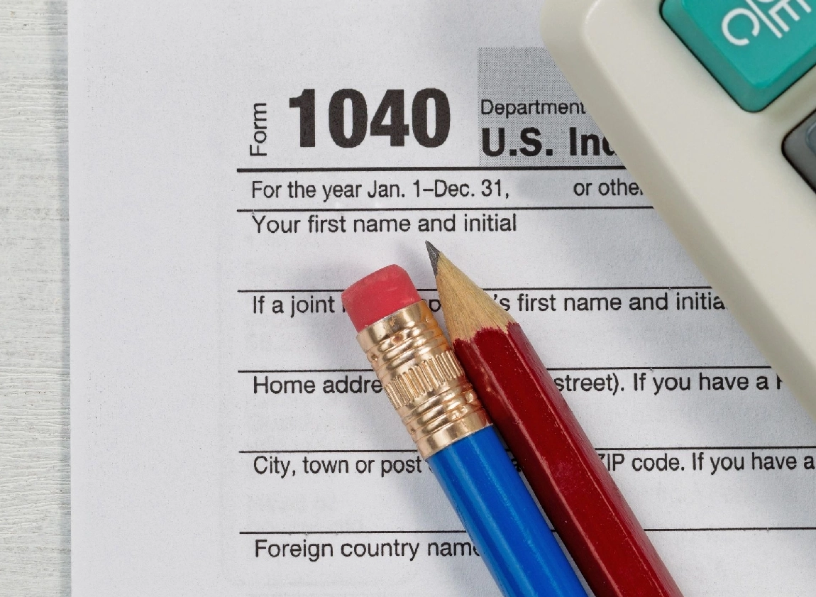 A 1040 tax form lies on a table next to a blue pencil, a red pencil with an eraser, and part of a calculator.