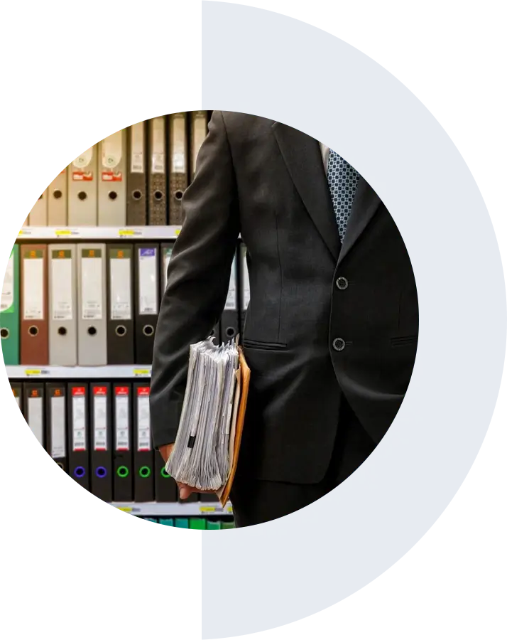 A person in a suit holding a stack of folders and documents in front of a shelf filled with various binders and files.