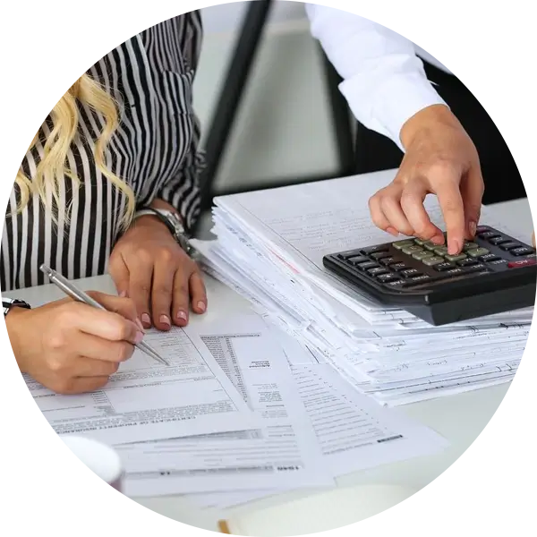 Two people are working at a desk, focusing on documents. One person is writing, and the other is using a calculator on top of a stack of papers.